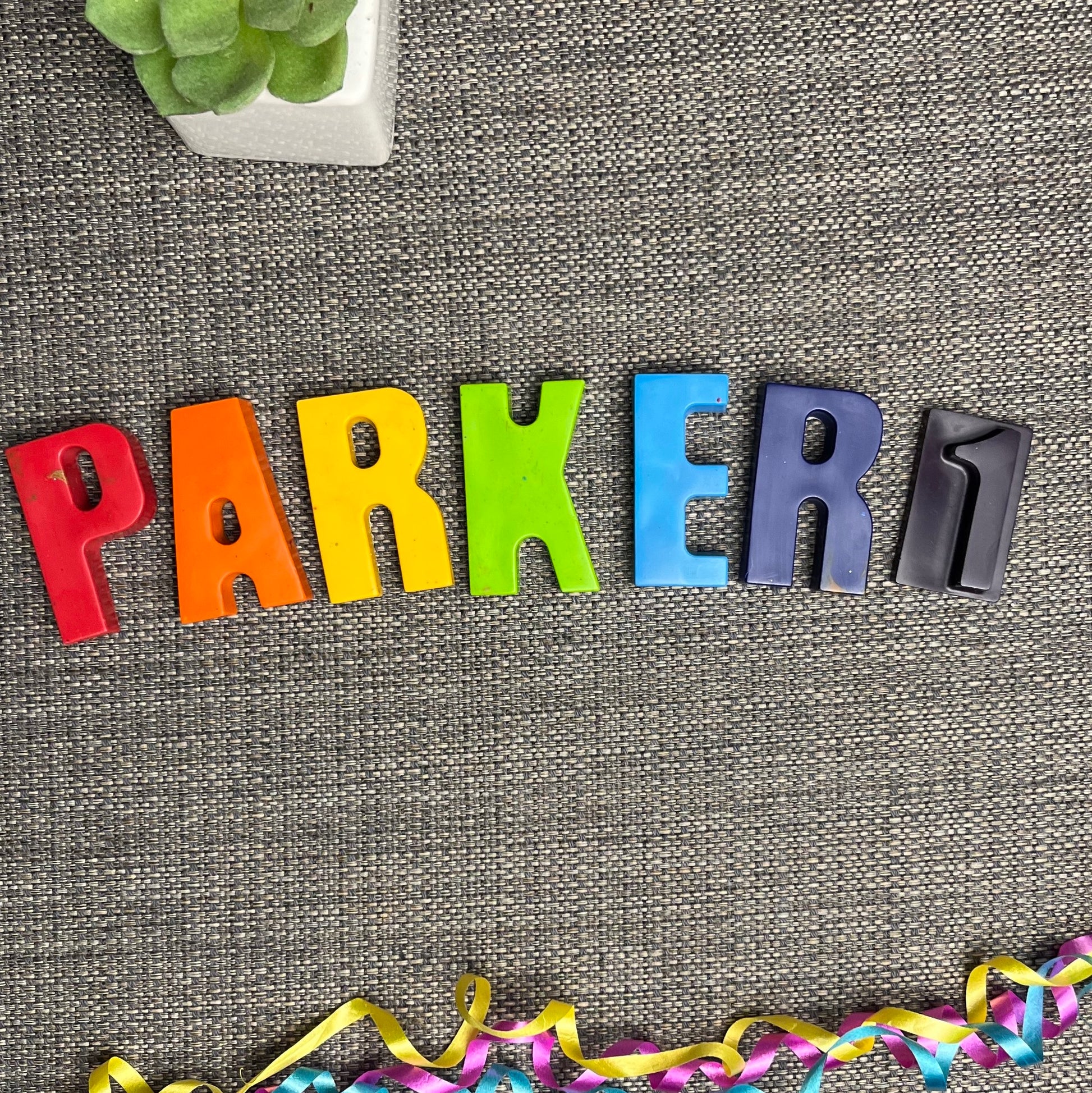 Letters for Parker1 made out of crayons with rainbow colours. P is red, A is orange, R is yellow, K is green, E is blue, R is dark blue and 1 is purple